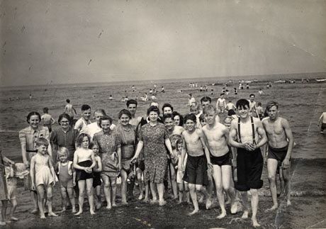 Photograph showing eight women in dresses, nine small children, seven young boys in swimming costumes, and four men, standing in a group in the shallows, on a beach with the sea behind them; many people can be seen swimming in the sea