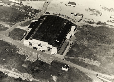 Photograph of an aerial view of a low building on its own with a road in front and to the side; at its back there appears to be the sands; the word Pavilion is written on its front; two vans and a car are parked in front of it