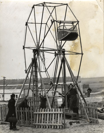 Photograph of four men erecting a ferris wheel with one carriage on it; a makeshift fence is round the wheel while they are working; behind them is a building which resembles The Pavilion at Crimdon Dene