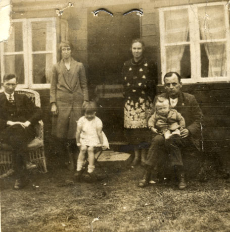 Photograph of two women standing either side of a door with a window on either side of it; two men are sitting either side of the women; one man is holding a small boy, aged approximately three years, and a small girl is sitting on a small table or stool between the two women; the men are in suits and one woman is in a coat and the other in a patterned dress; they have been identified as being at Crimdon Dene