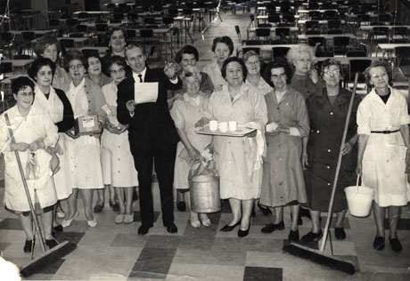 Photograph showing a group of sixteen women wearing overalls standing in what appears to be a canteen with tables and chairs behind them; a man in a suit is holding a piece of paper and gesturing; the photograph has been identified as Mr. T. Reynolds, Manager of Crimdon Park, directing the final touches to the Spring Cleaning of The Pavilion, Easter 1966