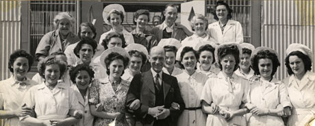 Photograph of twenty women in uniform and a man in overalls standing in a group with a man in a suit, who is smoking a cigarette; behind them is corrugated iron building possibly the canteen of Crimdon Park