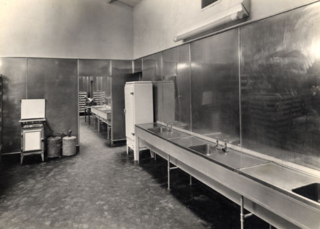 Photograph showing a line of stainless steel sinks at the right of the photograph with steel panels on the walls; on the left is a gas stove powered by bottled gas; further sinks and a meat cutter can be seen in a room beyond that nearer the camera; the rooms are part of The Pavilion at Crimdon Park, photographed for its opening; the photograph was taken by T. G. Reeves, 351 London Road, Mitcham, Surrey