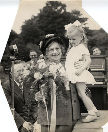 Photograph showing a middle-aged woman wearing a suit and hat embracing a small girl who is standing on a high surface near the woman who is holding in her right hand a bouquet of flowers, presumably presented to her by the girl; the girl, who is approximately six years of age, is wearing a short dress and has plaits; a man in a suit can be seen standing behind the woman next to a microphone; two other men can be seen in the distance with a mechanical piano