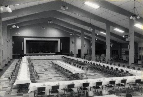 Photograph showing the interior of a large hall with a stage at the end and pillars along either side; tables have been arranged in a square running the length and breadth of the hall and set for a meal; the style of the architecture of the room is that of the 1950s