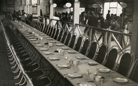 Photograph showing the surface of a long table laid with plates, knives and forks; the table is not the same as that shown in 28, because the backs of the chairs are different and the pillars in the hall are different; behind the table to the right of the picture people can be seen indistinctly near the self service counters