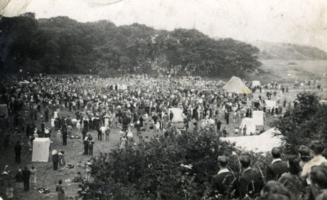 Photograph showing a large crowd of people, in the distance, on a field, with cliffs in the far distance; a marquee can be seen and what appear to be wind breaks; most of the people gathered on the field, however, are standing; a group of four young men and four young women can be seen from the rear watching the proceedings on the field at the right of the photograph
