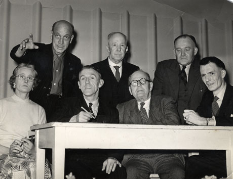 Photograph showing three elderly men and one elderly woman sitting at a wooden table; behind them three elderly men are standing; all but one of the men are dressed in suits and ties; the woman is dressed in a skirt and blouse; the woman and one of the men are holding pens; they have been identified as Pensioners' Rally at Crimdon