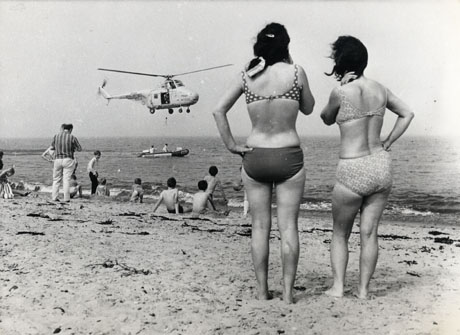 Photograph showing the rear of two young women wearing bikinis looking out to sea where an R.A.F. Rescue Helicopter, XP404, can be seen hovering above a Royal National Lifeboat Institute dinghy, in which there are two men; the rear of seven young boys, one man and a girl can be seen at the water's edge