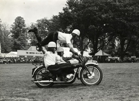 Photograph showing three young men wearing trousers, shirts, long boots and helmets performing tricks on a motor bicycle and side car; the registration number of the bicycle is HUE 557; the men are performing on an area of grass with crowds round the perimeter in the distance; a building with a sign reading The British Oil and Cake... can be seen behind the men; a sign reading Licensed Bar can be seen at the right of the picture; the photograph was taken by Graphic Photos, 411, St. Albans Road, Watford
