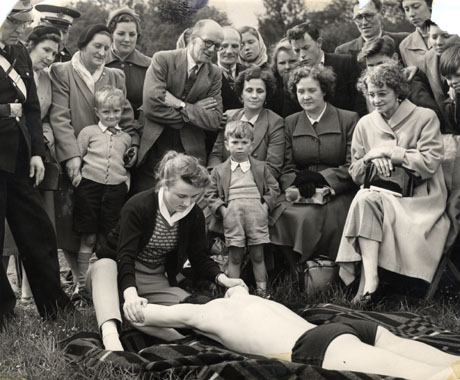 Photograph showing a young woman kneeling at the head of a young man wearing bathing trunks lying on his front on a rug; the woman is pulling his bent arms above his head; she is being watched by sixteen people and three children; also watching her are two men in uniform, presumably that of the St. John Ambulance Brigade; the photograph has been described as Crimdon Save a Life Demonstration