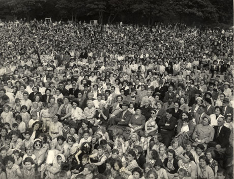 Photograph showing people sitting in rows completely covering the area of a field with trees behind them; children are sitting squashed together on the ground at the front of the photograph; the crowd is looking towards a spot just to the left of the camera; the photograph has been described as Bank Holiday Monday, 2nd August 1954