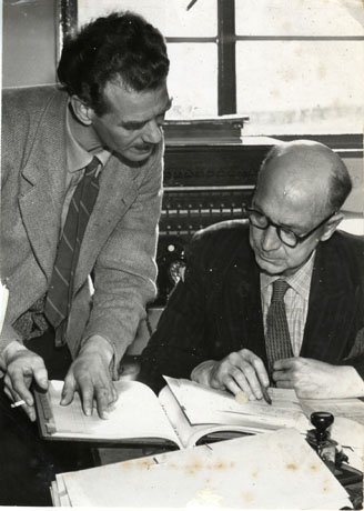 Photograph showing a man, dressed in a tie and tweed jacket and smoking a cigarette, standing at the left of the picture, next to a man, wearing a suit and tie, sitting at a desk; they are looking at a volume, possibly of accounts, and comparing the contents of the volume with a paper resting on the volume; a window and a pice of equipment can be seen behind them