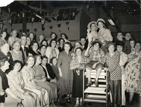 Photograph showing four rows of people sitting sideways to the camera at the left of the picture, looking at two women dressed as cowboys, one of whom is sitting on the end of a trolley; two women wearing straw hats are behind the woman on the trolley and three other women and three men are standing at the right of the trolley; it is possible that the individuals at the right of the picture have been entertaining those in the rows