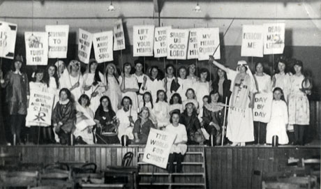 Photograph of approximately thirty one young people sitting and standing in a group on stage, dressed in costume possibly designed to reflect the history of The Book of Common Prayer, and carrying banners with exhortations to virtue; the photograph is described as Prayer Book Pageant, Deaf Hill