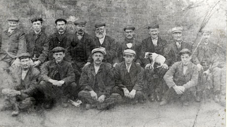 Photograph of a group of fourteen men in suits and caps sitting on the ground in front of brick wall; one of the men has a dog standing on his knees; the photograph is described as Meeting Place at Old Foundry, Deaf Hill