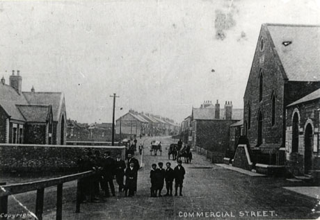 Postcard photograph entitled Commercial Street, and described as being at Deaf Hill; the photograph shows the street curving away into the distance to the right; on the right-hand side is the front of a non-conformist church and on the left-hand side a building which may be school; houses and shops can be seen indistinctly in the distance on the left-hand side of the street; a group of six men and six boys can be seen in the foreground and, beyond them, three horse-drawn carts are moving along the road