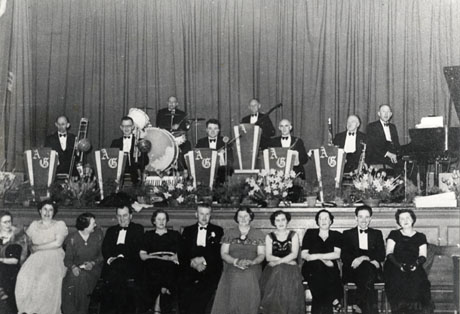 Photograph of eight members of a dance band sitting on a stage with stands in front of each player bearing the initials A G; below the stage a row of eight women and three men in evening dress are seated; the photograph is described as Mr. Grey's Band, Deaf Hill