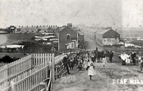 Postcard photograph entitled Deaf Hill, showing the ends of houses in a street curving away to the left; the gardens of the houses on the left of the street can be seen with washing on lines; in the foreground are groups of children and two or three carts; all the detail in the photograph is indistinct
