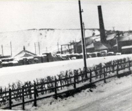 Photograph showing part of the surface of a road covered in ice with a low fence and hedge running alongside; beyond the fence is a field covered in snow, the roof of a building, a larger building with an industrial chimney and the pit heap, also covered in snow, in the distance; the photograph is described as being at Deaf Hill