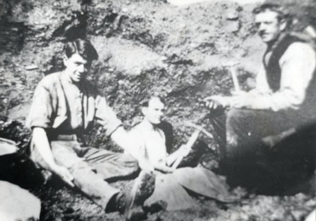 Photograph of three men, one of whom is holding a pick, and another a hammer, sitting against an indistinct background which may be a pit-heap; the photograph is described as Picking for Coal, 1922, Deaf Hill