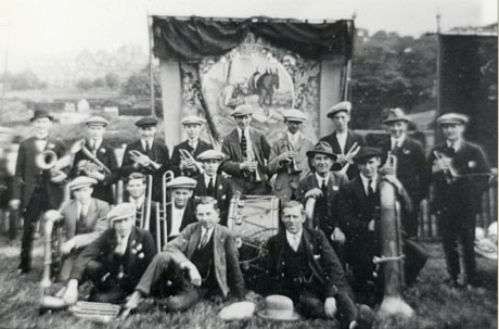 Photograph of a group of eighteen men of the Deaf Hill Welfare Band, posed in front of the Deaf Hill Lodge Miners' banner with their instruments on the ground in front of them; the banner shows a picture of the Good Samaritan; the photograph is a little blurred at the edges