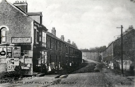 Postcard photograph entitled Station Lane, Deaf Hill, Trimdon Colliery, showing a street of terraced houses curving away to the left with the front of a shop at the end on the left; a group of three small indistinct figures can be seen outside the shop