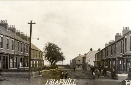 Postcard photograph entitled Deaf Hill, showing two rows of terraced houses with bay windows, standing opposite each other and separated by an un-made-up road; a number of small figures can be seen in front of the houses; the initials T W T can be seen in the corner of the photograph.