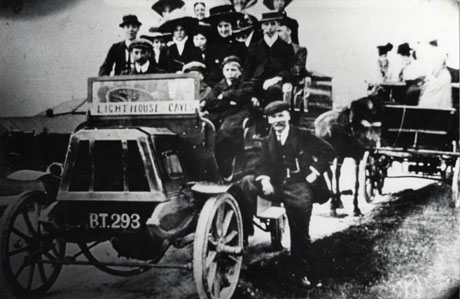 Photograph showing a group of fifteen people sitting in a motor charabanc with the registration B.T. 293 and a notice on its windscreen announcing Lighthouse Caves; behind the motor, a horse-drawn vehicle, with three or four indistinct figures in it, can be seen; the photograph is described as The Outing, Deaf hill
