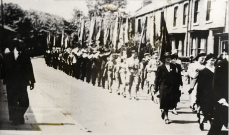 Photograph of a procession of people walking past terraced houses with bay windows; at the head of the procession are middle-aged women, followed by what appear to be Boy Scouts and, then, very indistinct ranks of individuals all carrying banners; the photograph is described as The Clock Procession, Deaf Hill
