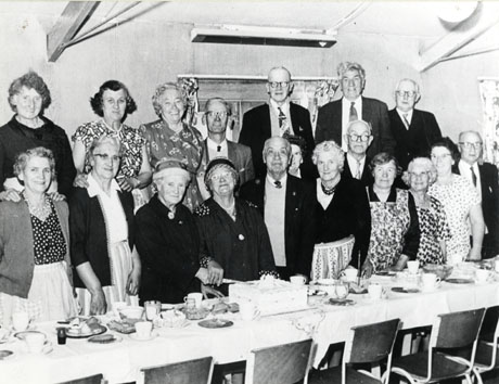 Photograph of a group of seven elderly men and twelve elderly women, standing behind a trestle table laid for tea in the Over-Sixties Club, and watching Mrs. Butterfield cutting a cake on the occasion of her birthday