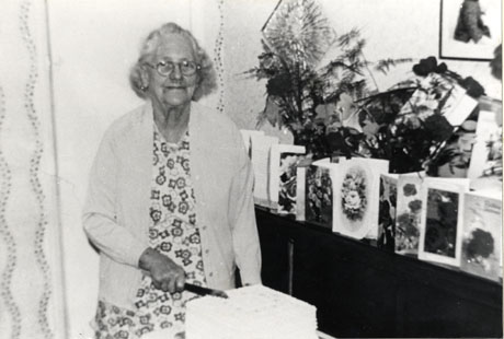 Photograph of Mrs. Butterfield standing to cut her cake on the occasion of her ninetieth birthday, next to a sideboard containing cards and flowers at Deaf Hill