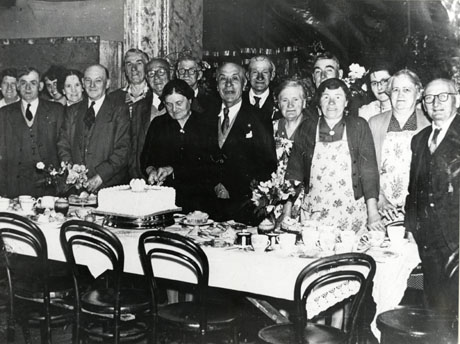 Photograph of a group of nine men and eight women standing behind a table laid for tea and watching a woman and a man cutting a cake on the occasion of the Over Sixties Christmas Party at Deaf Hill