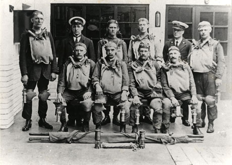 Photograph of the eight members of the Rescue Brigade, Deaf Hill Colliery, posed wearing their rescue equipment and carrying their miners' lamps; a stretcher is rolled up on the ground in front of the group
