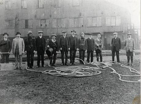 Photograph of twelve men, described as the Deaf Hill Rescue Brigade, posed outside what appears to be colliery buildings; all of the men are wearing suits, but two of them are wearing helmets, one is carrying a pair of bellows which are attached to the end of a length of tubing which is spread out in front of the group, and two others are holding equipment