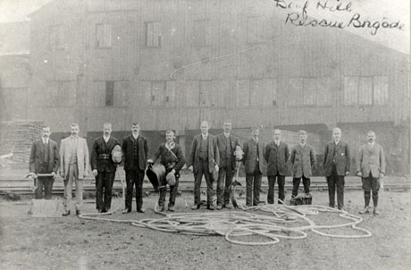 Photograph of the twelve members of the Deaf Hill Rescue Brigade, posed outside what appear to be colliery buildings, on the same occasion as in deaf0030; the photograph is taken from a slightly longer distance and the members of the group have removed their hats and helmets