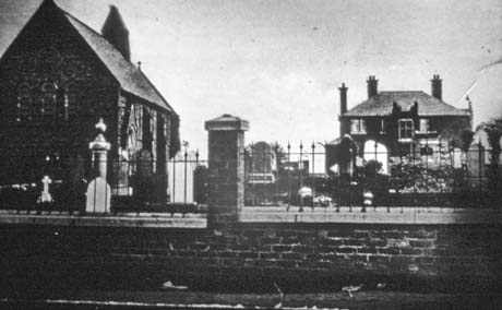 Photograph showing the wall of a graveyard with railings on top of it; beyond the wall, grave stones and monuments can be seen; at the left of the picture, the end and side of a church can be seen indistinctly; beyond the graveyard, the front of a house with high chimney stacks can be seen; the photograph has been identified as Deaf Hill Church and Vicarage