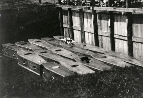 Photograph of fifteen coffins lying in the mass grave of the victims of the colliery disaster at Easington Colliery in 1951; an official cap is lying on one of the coffins and a Union Jack with what appears to be a black circle in the middle of it on another