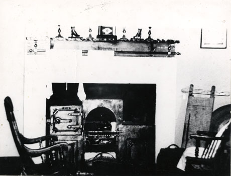 Photograph of a kitchen range showing its fireplace, oven and mantleshelf above with ornaments; in front of the range is rocking chair and a Windsor chair; the photograph is described as Interior of Typical Colliery House