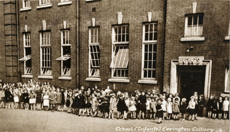 Postcard photograph entitled School, (Infants) Easington Colliiery.4, showing children standing in the playground outside the school with the windows of the school and the entrance marked Infants behind them