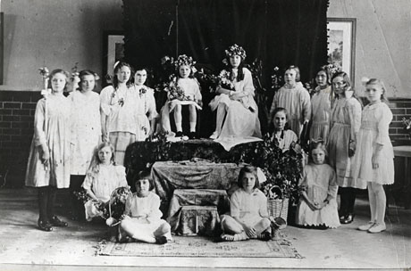 Photograph of fifteen girls, between the ages of approximately seven and eighteen years, wearing light coloured dresses and grouped round and sitting on a dais inside a building; the girls are holding branches of flowers and two are wearing wreaths of flowers; the photograph is described as May Day Celebrations at Easington Girls' School, 1925