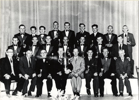 Photograph of twenty four men, most of whom are in evening dress, and holding small trophy cups, posed against a curtain; the man in the middle of the front row is holding a large trophy; the photograph is described as Easington Public Band