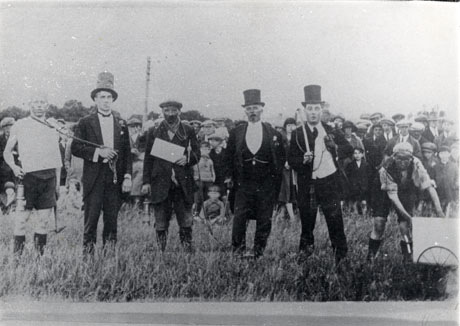 Photograph described as Carnival, Easington Colliery, showing a large group of people in a field with six men in the foreground in fancy dress; three of these are dressed as miners and three are dressed in evening clothes and top hats; one of the figures in evening dress has a noose round the neck of one of the miners