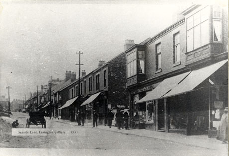 Postcard photograph entitled Seaside Lane, Easington Colliery. 1330 showing one side of a street of shops going uphill; the awnings are out on most of the shops and a cart can be seen from the rear; figures can be seen on the pavement