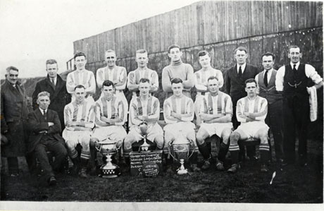 Photograph of the eleven members of Easington Athletic Football Club, posed against a fence with six men in suits; three trophies are on the ground in front of the team; one is standing on a box on which is written: Trophies Won. Easington W. and Wearside League Champs. National Orphanage Cup Durham.....; on a ball in front of the box is written: 1931-32 Winners Wearside League Durham ........