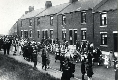Photograph showing one side with terraced houses of Station Road, Easington Colliery, with a procession of children walking down the road; a large number of people can be seen walking behind the procession and watching from the pavements; the photograph is described as Easington Colliery Carnival, Station Road, 1930