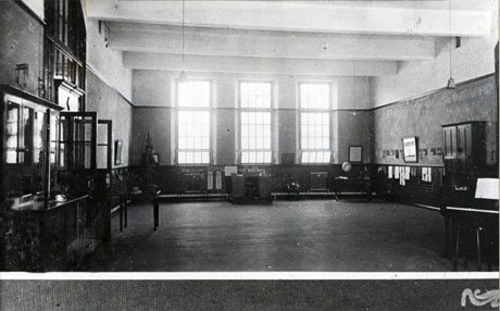 Photograph of the interior of a school hall with cupboards, tables, a piano, indistinct pictures on the walls, a Union Jack and long windows at Easington Junior Mixed School