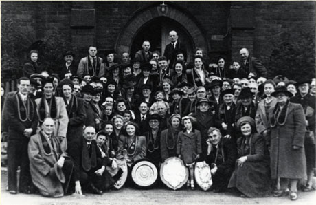 Photograph of a large group of approximately a hundred men, women and children, posed outside a large building; each of the adults wears a badge round his or her neck; pieces of silver are displayed at the front of the group; the photograph is described as Good Templars, Easington