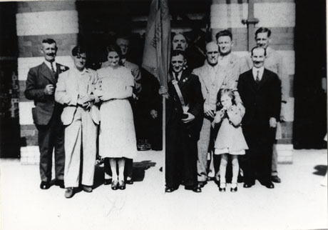 Photograph of a group of nine men, one woman, and one child, posed outside a large building, in Easington Colliery, with contrasting bands of different coloured brick; one man is holding a British Legion Flag