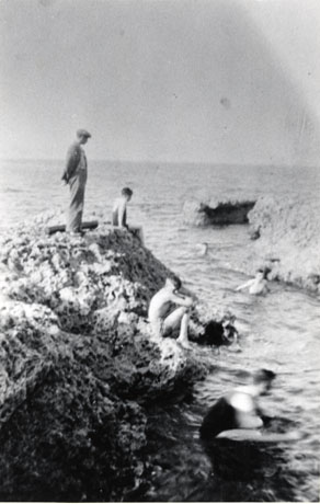 Photograph showing a man, wearing a jacket and cap, standing on a rocky promontory with the sea on both sides, and a boy swimming in the sea and two boys sitting on the rock near the man; a blurred figure is in the sea at the front of the picture; the photograph has been identified as Easington Colliery Beach